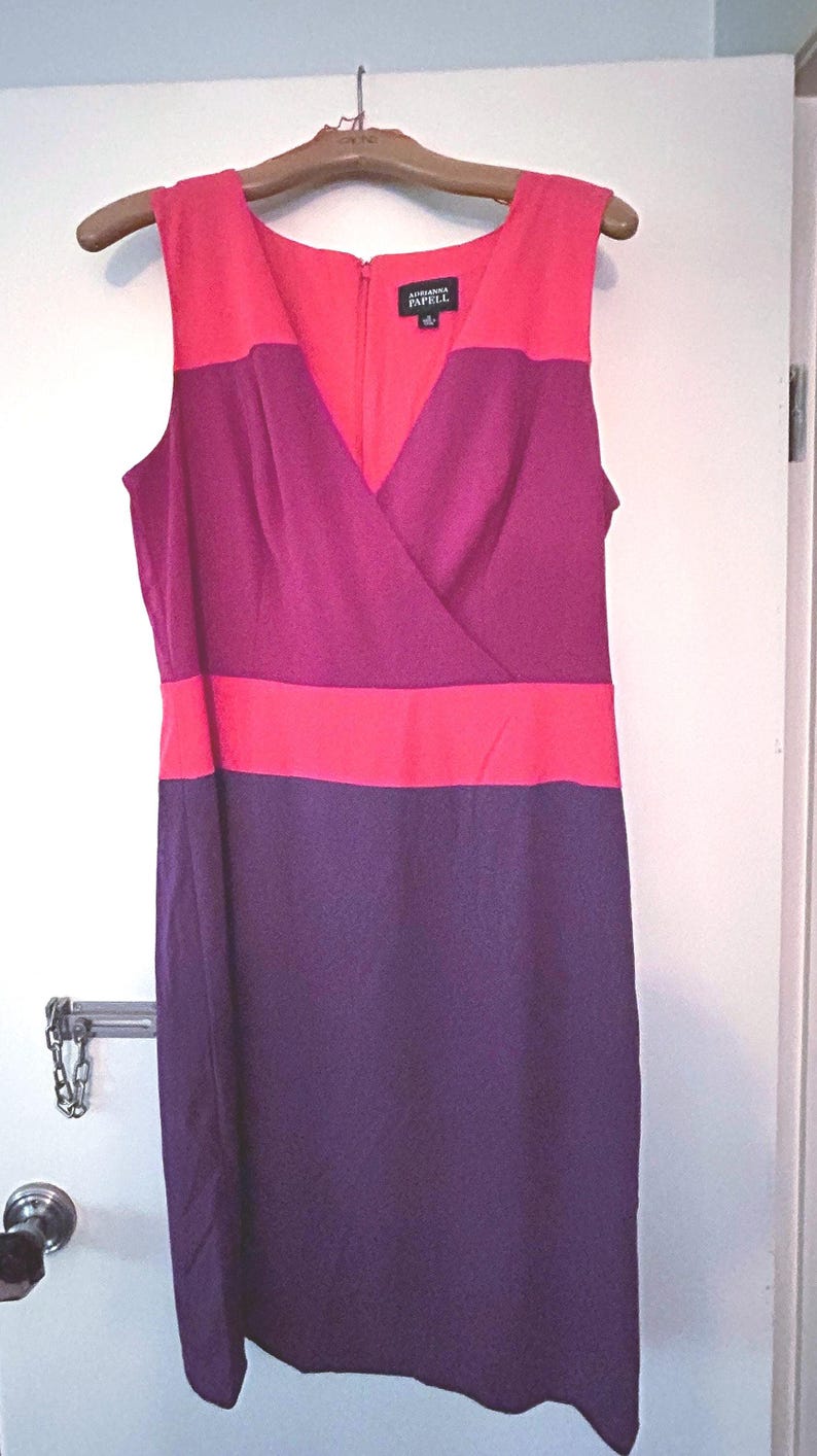 May include: Sleeveless color block dress in shades of red, fuchsia, and purple. The dress features a V-neckline and a knee-length hem. The brand name "Adrianna Papell" is visible on a tag near the neckline. The dress is hanging on a wooden hanger.