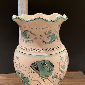 May include: A white ceramic vase with a scalloped rim and a green and black floral design. The vase features a painted portrait of a woman with a green hat.