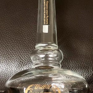 May include: A clear glass Willett Bourbon Whiskey bottle with a gold label. The label reads "Kentucky Bourbon Whiskey" and "94 Proof." The bottle has a long neck and a wooden stopper. The text "Small Batch Bourbon made in Kentucky" is printed on the neck.
