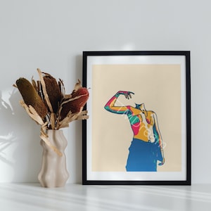 Puede incluir: Una impresión artística enmarcada con una figura abstracta colorida sobre un fondo crema. La figura se representa con tonos vibrantes de azul, amarillo, rosa y verde. La obra de arte se exhibe sobre una superficie blanca con un jarrón de flores secas.