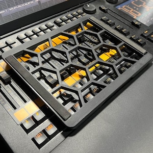 May include: A black honeycomb-patterned overlay rests on a mixing console, featuring buttons, sliders, and a digital display. The overlay's design exposes the console's orange-lit keys and controls. The image provides a close-up view of the equipment.
