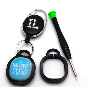 Può includere: Una collezione di accessori, tra cui un portachiavi nero con le lettere "IL", un portachiavi con la scritta "THE HAPPIEST PLACE ON EARTH", un anello nero e un piccolo cacciavite con la parte superiore verde.