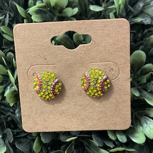 May include: A pair of stud earrings featuring a softball design with yellow and red rhinestones.