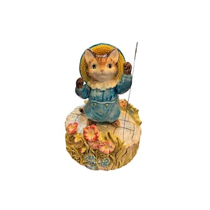 Puede incluir: Figura de cerámica de un gato con abrigo azul y sombrero amarillo, sosteniendo una caña de pescar. El gato está sobre una base decorativa con flores. La figura tiene una estética vintage y caprichosa.