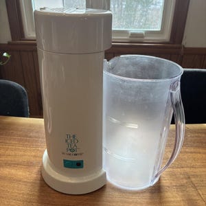May include: White "The Iced Tea Pot by Mr. Coffee" appliance with a clear plastic pitcher. The tea maker has a cylindrical design with a rounded top and a teal power button. The pitcher has a handle and measurement markings.