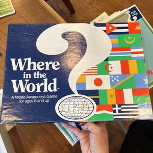 May include: A rectangular board game box titled Where in the World, A World Awareness Game for ages 8 and up. The left side is dark blue with a large white question mark and a globe icon. The right side features a colorful grid of national flags.