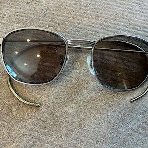 May include: Silver-framed sunglasses with dark lenses. The round lenses are set in a silver frame with a thin bridge and temples. The temples have a black and gold striped design. The sunglasses are resting on a textured, gray surface.