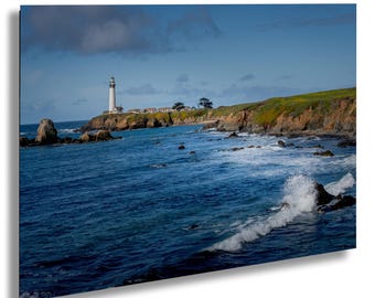 May include: A tall white lighthouse stands on a grassy cliff overlooking a deep blue ocean with crashing waves. The coastline features rocky formations and a clear blue sky with scattered clouds above the horizon on a bright sunny day.