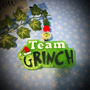 May include: A green Christmas ornament with the words "Team GRINCH" in white and black. The ornament has a red Santa hat and is decorated with a Grinch face. The ornament is hanging from a string of beads and artificial leaves.