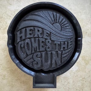 May include: A black, circular ashtray with the phrase "HERE COMES THE SUN" in stylized lettering. The design includes a sun and wave graphic. The ashtray has a rectangular notch and is set against a neutral background.