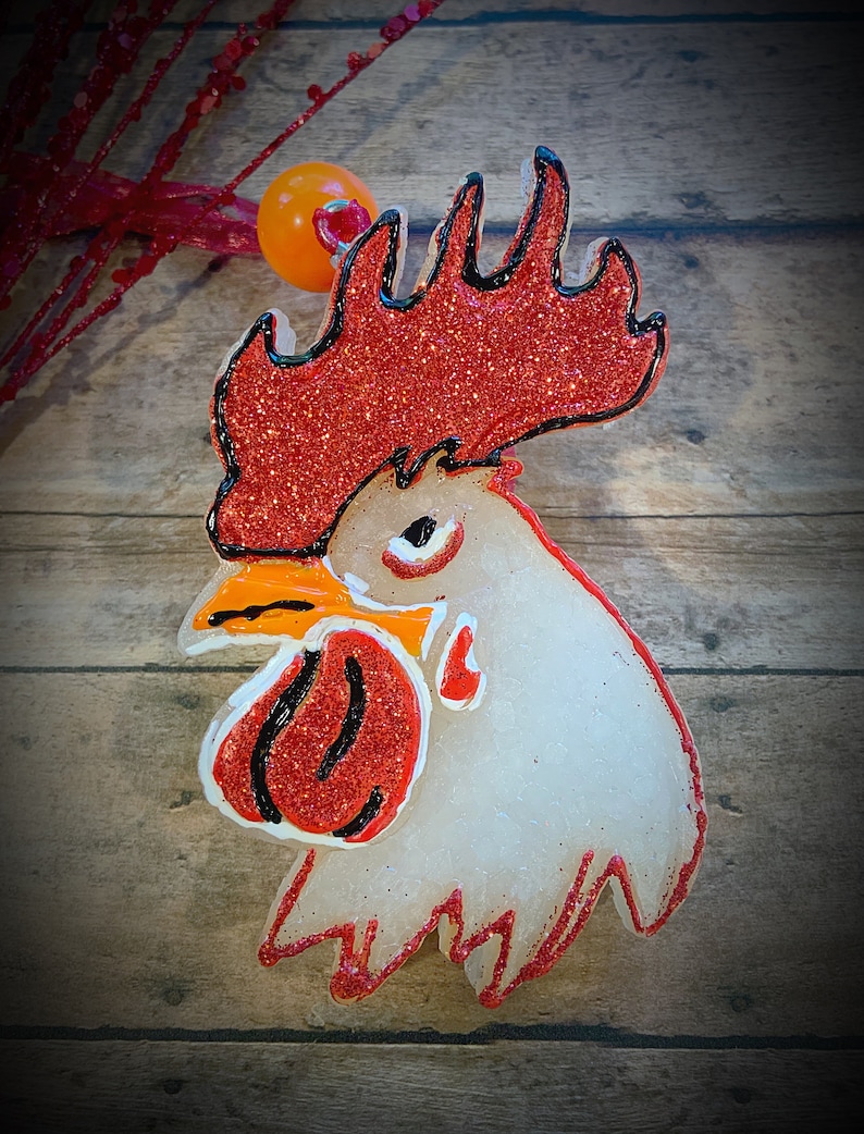 May include: A decorative rooster head ornament with a red glitter comb and wattle, outlined in black. The rooster's head is white with an orange beak. The ornament is set against a wooden background.