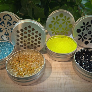 May include: Six round metal containers with different designs on the lids. Each container is filled with colorful beads. The beads are in shades of blue, yellow, gold, and black.