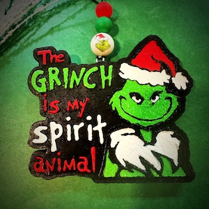 May include: A Grinch-themed ornament with the text "The Grinch is my spirit animal." The Grinch is depicted in green with a Santa hat. The ornament has a black border and is accented with red, green, and white beads.