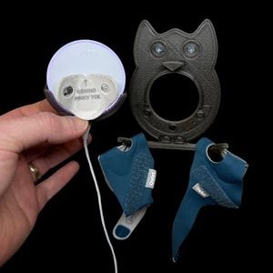May include: A baby monitor system with a round white sensor, a dark gray owl-shaped base, and two blue fabric socks. The sensor has the text "Owlet" and "Behind Pinky Toe". The owl base has two blue gems for eyes.