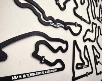 May include: A collection of black, textured wall-mounted racing circuit outlines. The tracks include the Miami International Autodrome and Jeddah Corniche, each featuring white text labels and small checkered finish line markers on the track paths.
