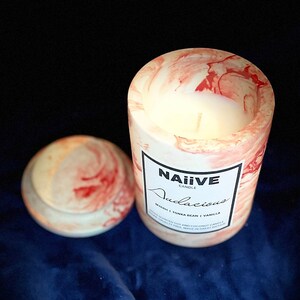 May include: A white candle with a red and white marbled design in a glass jar. The jar has a label that reads "NAÏVE CANDLE Audacious MYRRH | TONKA BEAN | VANILLA HAND-SCENTED SOY AND COCONUT CANDLE PARABEN FREE | MADE IN GREAT BRITAIN"