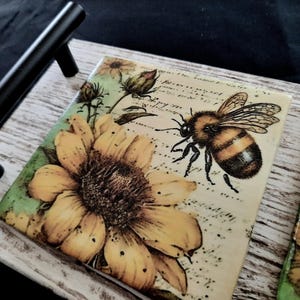 May include: A decorative tray with a distressed white wooden frame and black metal handles. The tray features two square ceramic tiles with a yellow sunflower and a bee illustration. The tiles have a vintage, botanical theme.