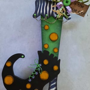 May include: A decorative wooden cutout shaped like a curled-toe shoe, featuring a green shaft with orange polka dots and a black foot section with orange dots. It includes a striped cuff, a green button, ribbons, and a tag reading if the shoe fits.