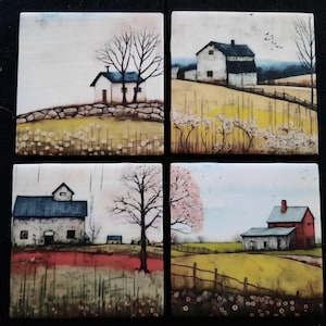 May include: Set of four square ceramic coasters, each featuring a different watercolor painting of a rural landscape. The paintings depict white houses, trees, and fields in various seasons. The coasters are set against a black background.
