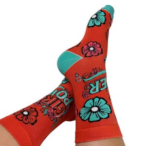 May include: A pair of orange socks with a floral pattern and the words "GIRL POWER" in a stylized font. The socks have teal accents on the toes and heels, and feature pink and teal flowers.