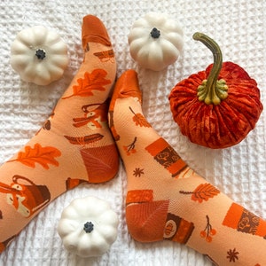 May include: A pair of orange and white socks with a fall-themed design featuring leaves, pumpkins, and a coffee cup. The socks are on a white fuzzy surface with small white pumpkins and a large orange velvet pumpkin.