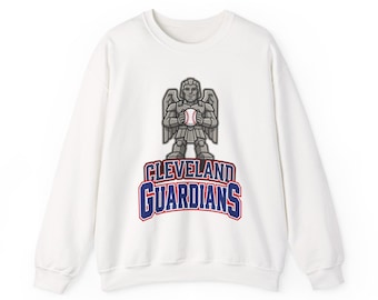 May include: A white long-sleeve crewneck sweatshirt featuring a graphic of a gray stone-like winged guardian figure holding a baseball. Below the figure, the text CLEVELAND GUARDIANS is printed in bold blue letters with a red outline.