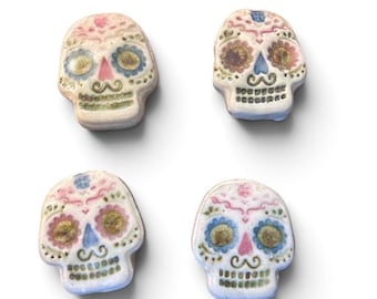 May include: Four ceramic skull beads featuring hand-painted details in pastel pink, blue, and green. Each skull displays a decorative floral pattern around the eye sockets, a small triangular nose, and a mustache-like design above the teeth.