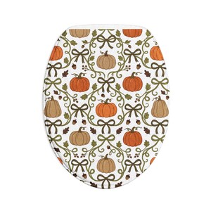 Op de afbeelding: Een ovale toiletbrilhoes met een witte achtergrond en een herhalend patroon van oranje en beige pompoenen, donkergroene krullende ranken, bruine eikenbladeren, kleine eikeltjes en donkergroene strikken in een symmetrische opstelling.