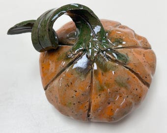 May include: A glazed ceramic pumpkin sculpture featuring a glossy orange body with dark speckles and deep vertical grooves. The top includes a prominent, curved, dark green stem with a leaf-like extension, all finished with a reflective surface.
