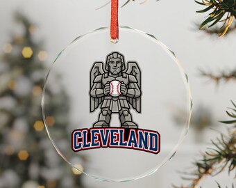 May include: A clear, circular glass ornament with a red ribbon hanger. The center features a graphic of a gray, winged, armored figure holding a baseball. Below the figure, the word CLEVELAND is printed in bold, blue, block letters.