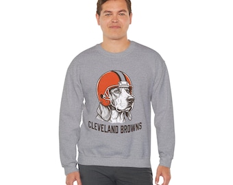 May include: A heather gray long-sleeve sweatshirt featuring a graphic of a hound dog wearing an orange football helmet with a brown and white stripe. Below the dog, the text CLEVELAND BROWNS is printed in a dark brown, bold, sans-serif font.