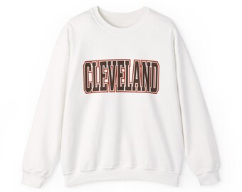 May include: A white long-sleeve crewneck sweatshirt featuring the word CLEVELAND printed across the chest in dark brown block lettering with a thin red outline. The garment has a ribbed collar, cuffs, and hem, providing a clean look.