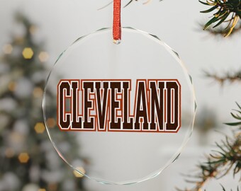 May include: A clear, circular, faceted glass ornament featuring the word CLEVELAND printed in bold, dark brown block letters with a thin orange outline. The ornament hangs from a thin, vibrant red ribbon loop at the top center.