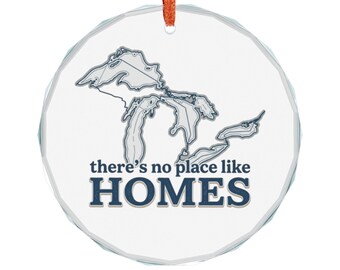 May include: A circular, faceted glass ornament featuring a blue outline map of the Great Lakes. Below the map, the text reads there's no place like HOMES in blue lettering. The ornament hangs from a thin, bright orange ribbon loop.