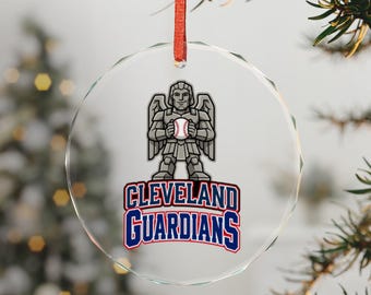 May include: A circular clear glass ornament with a red ribbon hanger. The center features a gray stone-like winged guardian figure holding a baseball, with the text CLEVELAND GUARDIANS printed in bold blue and red lettering below it.