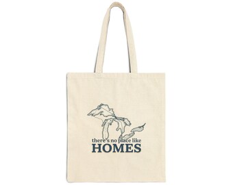 May include: A beige canvas tote bag featuring a dark blue line art illustration of the Great Lakes. Below the map, the text reads there's no place like HOMES in a combination of lowercase and bold uppercase lettering centered on the bag.