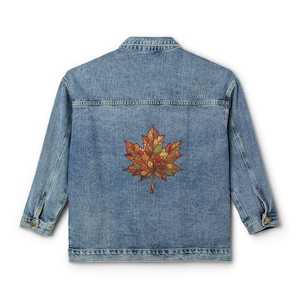 Könnte beinhalten: Eine mittelblaue Jeansjacke mit langen Ärmeln, Kragen und geknöpften Manschetten. Auf der Rückseite der Jacke befindet sich eine große, detaillierte Grafik eines einzelnen Ahornblatts, das aus verschiedenen herbstlich gefärbten Blattmustern besteht.