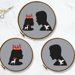 May include: Three cross-stitch embroidery hoops with black silhouettes. One hoop features a woman with flames atop her head and a deer, facing a man with a bird on his shoulder. The other two hoops show similar designs.
