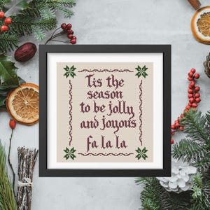 Puede incluir: Una obra de arte en punto de cruz enmarcada con el texto "Tis the season to be jolly and joyous fa la la" en hilo burdeos. La obra de arte está en un marco negro, rodeada de vegetación navideña, bayas y rodajas de naranja secas, creando un tema festivo.