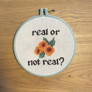 May include: A white embroidery hoop with a blue border, featuring a cross-stitch design of three orange flowers with green leaves and the text "real or not real?"