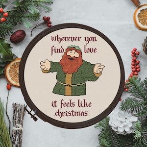 Peut inclure: Une broderie au point de croix ronde et encadrée représentant un personnage de dessin animé avec une barbe rousse et un manteau vert. Le texte dit "où que vous trouviez l'amour, c'est Noël".