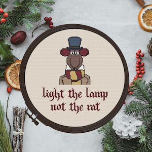 Può includere: Arte ricamata a forma di cerchio con un ratto dei cartoni animati che indossa un cappello a cilindro e una sciarpa. Il testo "light the lamp not the rat" è ricamato con filo rosso.