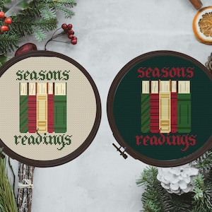 May include: Two round embroidery hoops with a green and red cross-stitch design of stacked books. The text "seasons readings" is stitched in black on a cream background in one hoop and a dark green background in the other hoop.