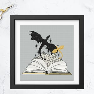 May include: Framed cross-stitch art featuring a black dragon soaring above an open book with a gold dragon and clouds. The artwork is in a black frame, with gold scissors in the upper left corner. The background is a light gray.