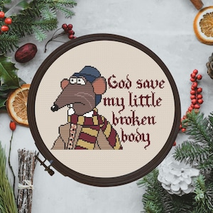 May include: A circular embroidery hoop containing a cross-stitch design of a brown rat wearing a blue hat and a yellow and red striped scarf. The text "God save my little broken body" is stitched in dark red letters to the right of the rat.