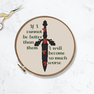 May include: A cross-stitch embroidery hoop with a black sword design and the text "If I cannot be better than them I will become so much worse" in green. Gold scissors are in the upper left corner.