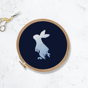 以下が含まれることがあります： 暗い青色の布地に白いウサギのシルエットが刺繍され、木製の刺繍枠に張られています