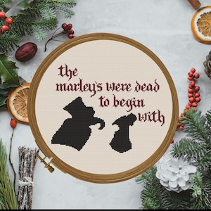 May include: A cross-stitch embroidery hoop with the text "the marley's were dead to begin with" and two black silhouettes. The embroidery is set against a cream-colored fabric, framed by a wooden hoop. Decorative elements include pine branches and dried orange slices.