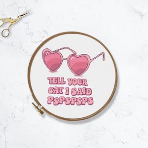 Puede incluir: Un aro de bordado blanco con gafas de sol de corazón rosa y el texto "TELL YOUR CAT I SAID PSPSPSPS".