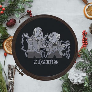 May include: A circular embroidery hoop containing a black fabric piece with a cross-stitch design of two elderly men in suits wrapped in chains. The text CHAINS is stitched in white below the figures. The hoop has a dark brown frame.